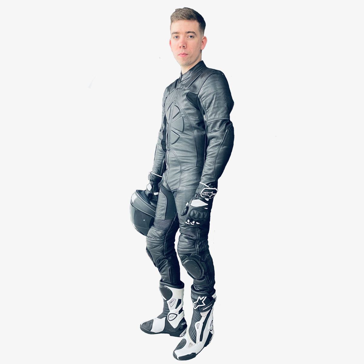 Man wearing a full-body black leather motorcycle racing suit holding a helmet on a white background - Side view