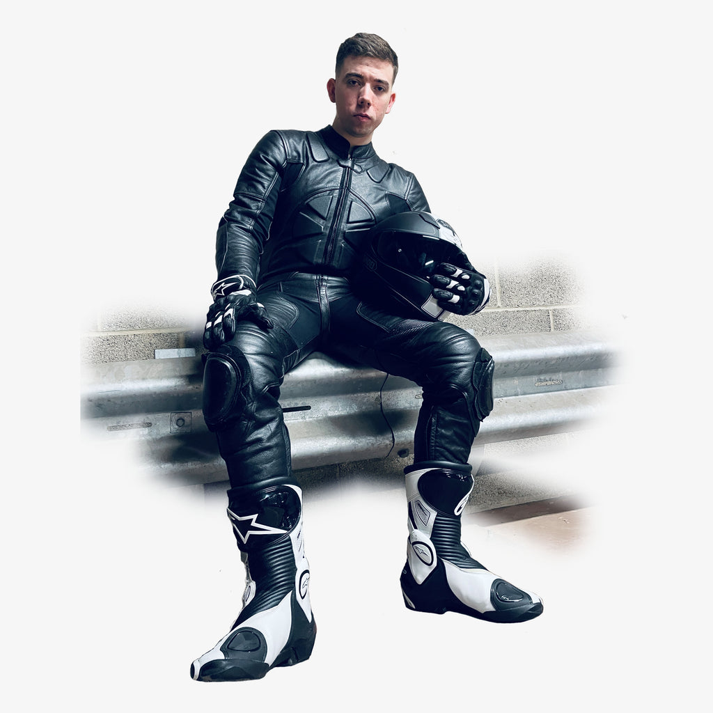 Man wearing a full-body black leather motorcycle racing suit holding a helmet on a white background - Sitting down